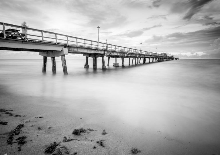 Black-and-white view of a long pier above smooth surf, with a blurred person, cloudy sky, and seaweed on the sand.