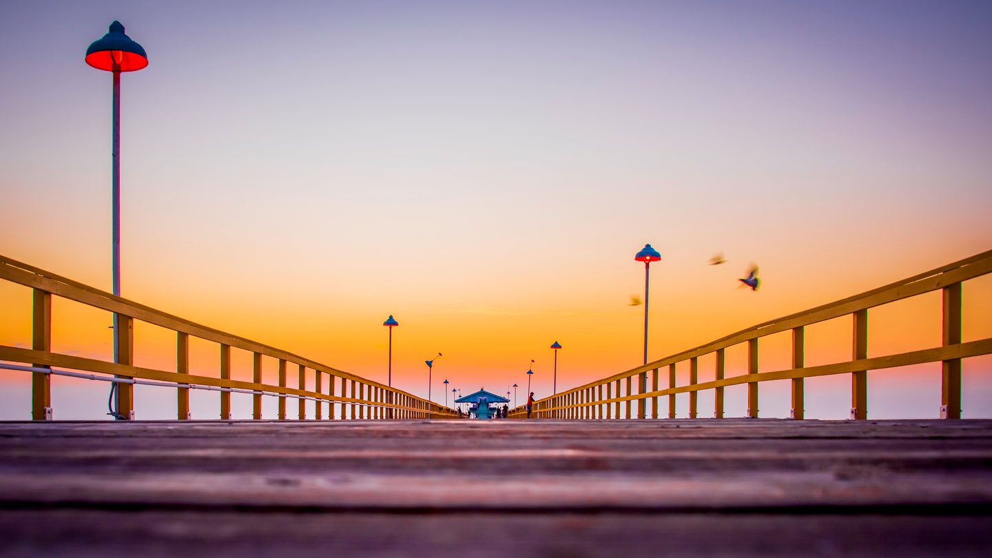 Low view along a wooden pier with yellow railings, glowing lamps, distant people, and birds against an orange sky.